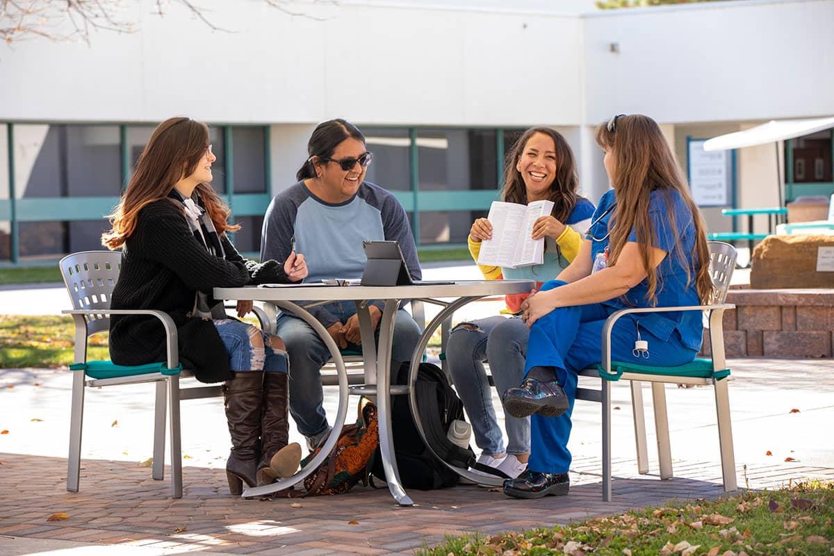 四个太阳集团娱乐场登陆网站的学生坐在外面的一张桌子旁交谈和微笑.