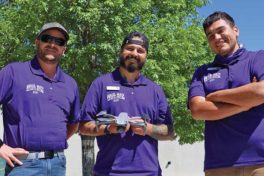 Three men wearing purple shirts standing holding a drone