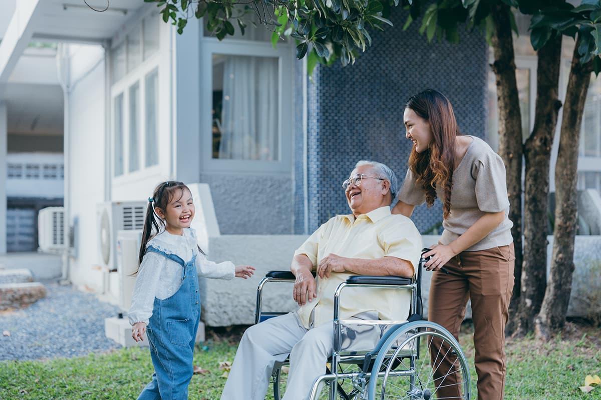 社会工作者 laughs along with a wheelchair bound elderly man 和 his gr和aughter outside a house under a tree.