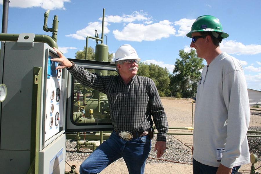 Two individuals in hard hats look at each other while standing next to a natural gas meter